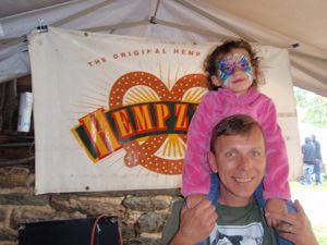 Man holding face painted young girl under tent in front of Hempzel Pretzel Sign at Fairy Fest.