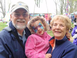 Grandparents on either side wearing blue man wearing cap holding facepainted child in pink top outdoors.