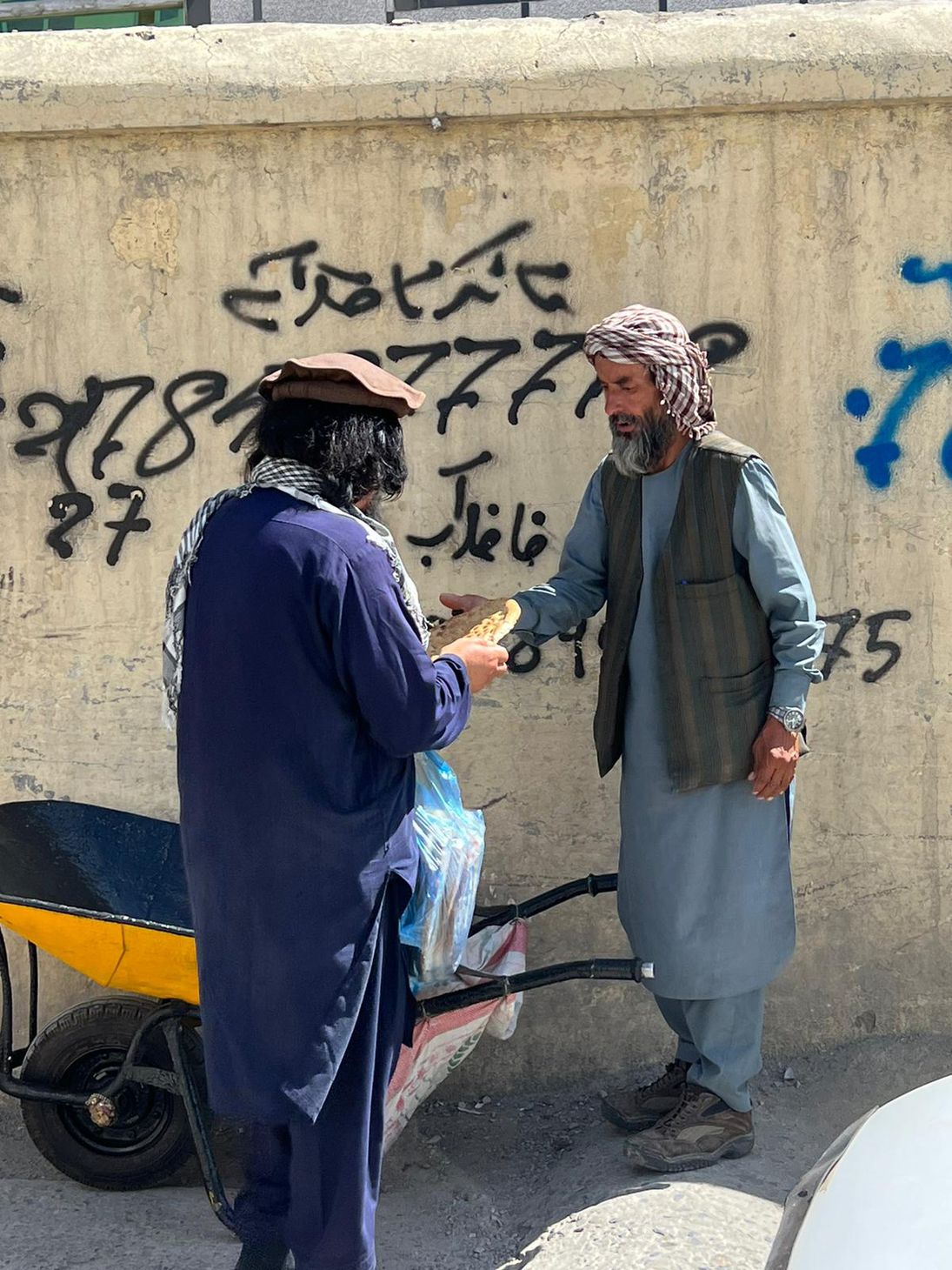 Two men one in purple hands hemp bread to man in blue background wall has Afghanistan grafiti.