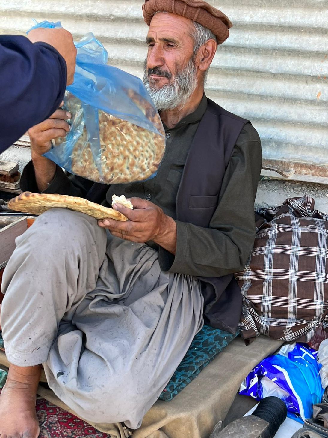 Old man without shoes dressed in blue with turban sitting on sidewalk and eating the hemp bread.