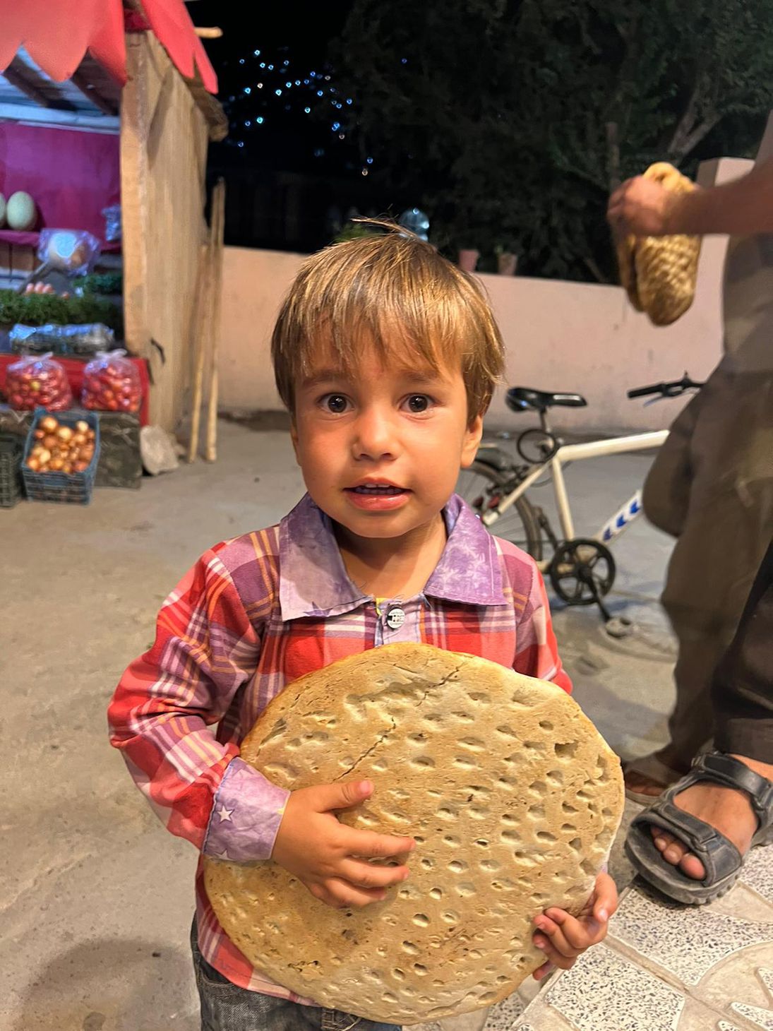 Blond headed little boy in red flannel holds close to his chest a big piece of hemp bread with a big smile on his face out in the market with bikes, stands.