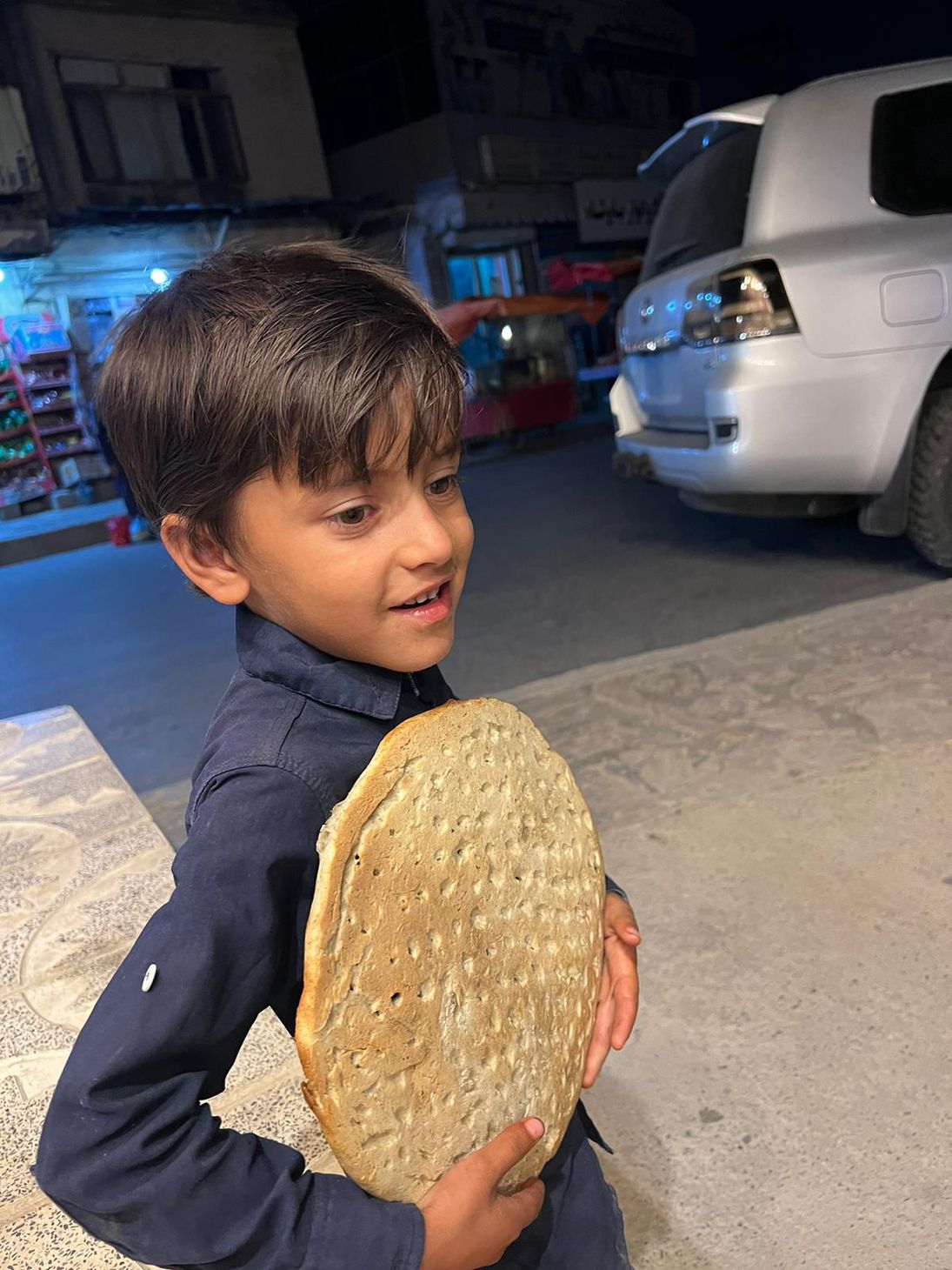 Little black haired boy wearing dark blue button up shirt smiles and clutches his hemp bread next to a car in the background.