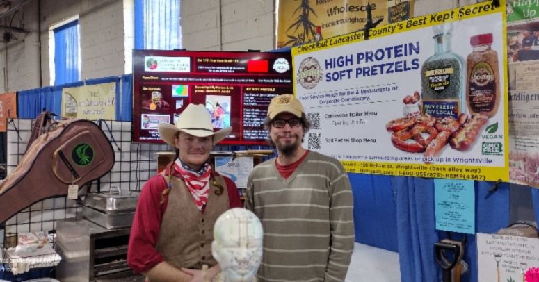 Two volunteers in Hempzels(tm) booth one wearing cowboy had & American flag kerchief and vest and other in baseball cap & dune color hemp sweater with TV in background.