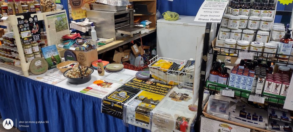 Merchandise on a long table with blue skirting, pretzels, black and gold bag and racks of products.