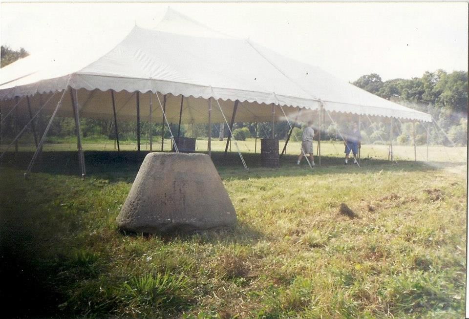 A hemp millstone sits on a grass field with an empty large tent with 3 people setting up underneath it.