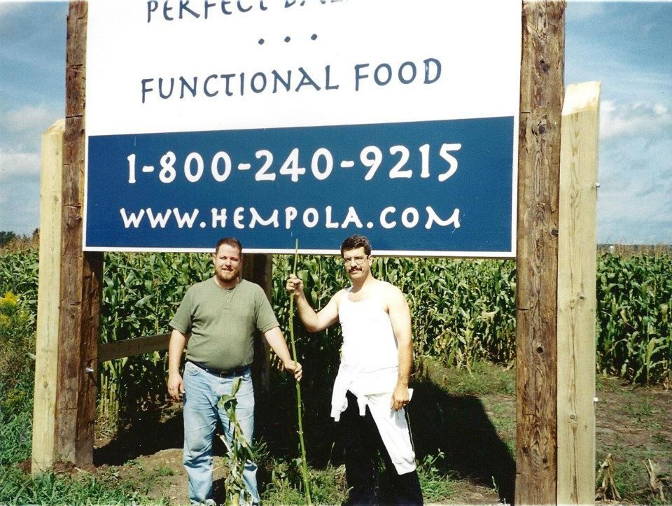 Jim Stark in green hemp tshirt with Shawn house in white tank top holding hemp fiber under giant Canadian Hempola Farms Wooden Sign near cornfield.