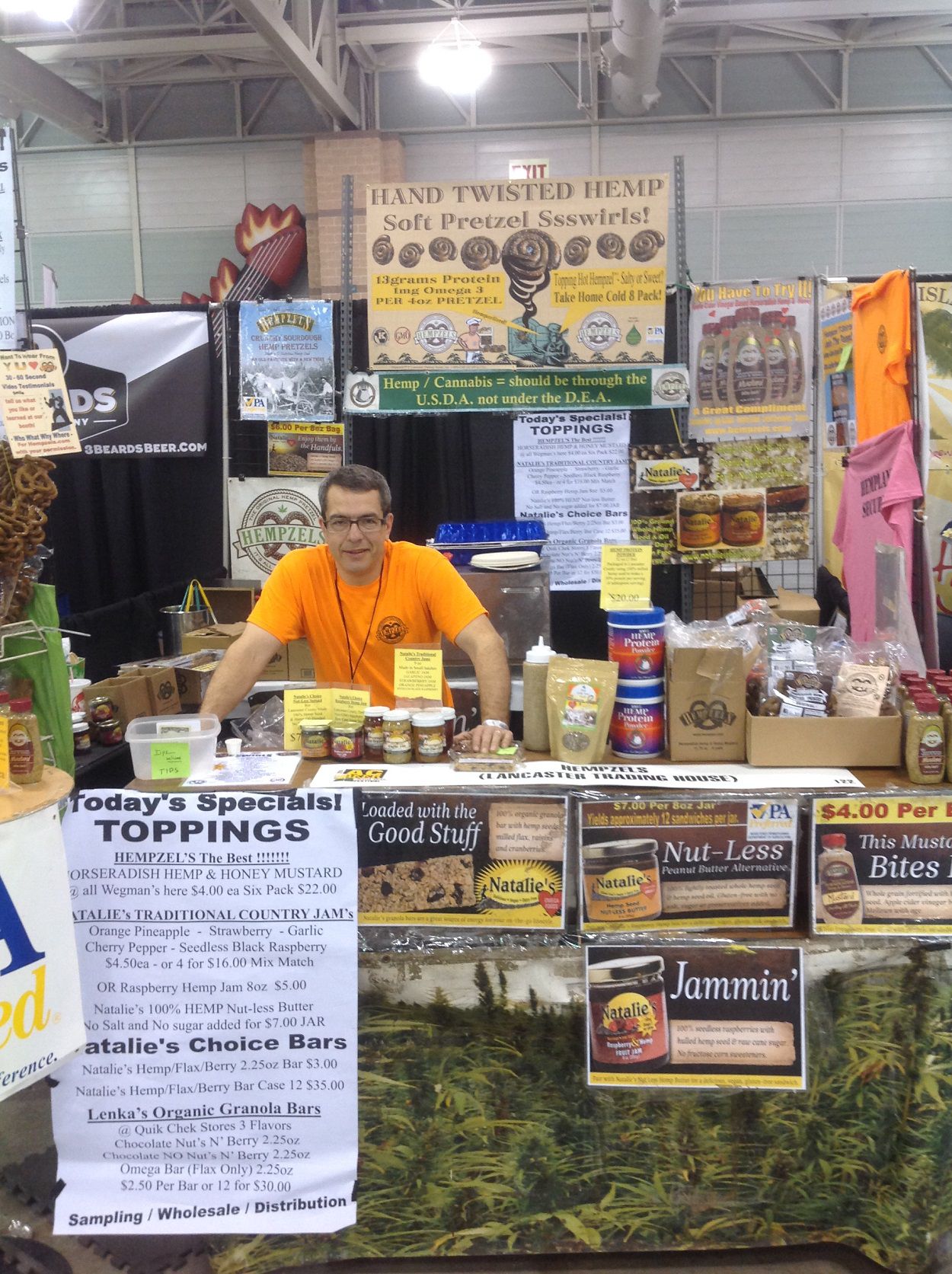 Owner Shawn in orange Hempzels(tm) tshirt with both hands on the counter in the booth with signs and products for sale.