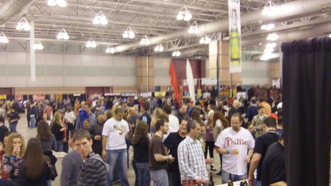 Big crowd inside a giant convention hall with bright lights and colorful flags some holding beer.