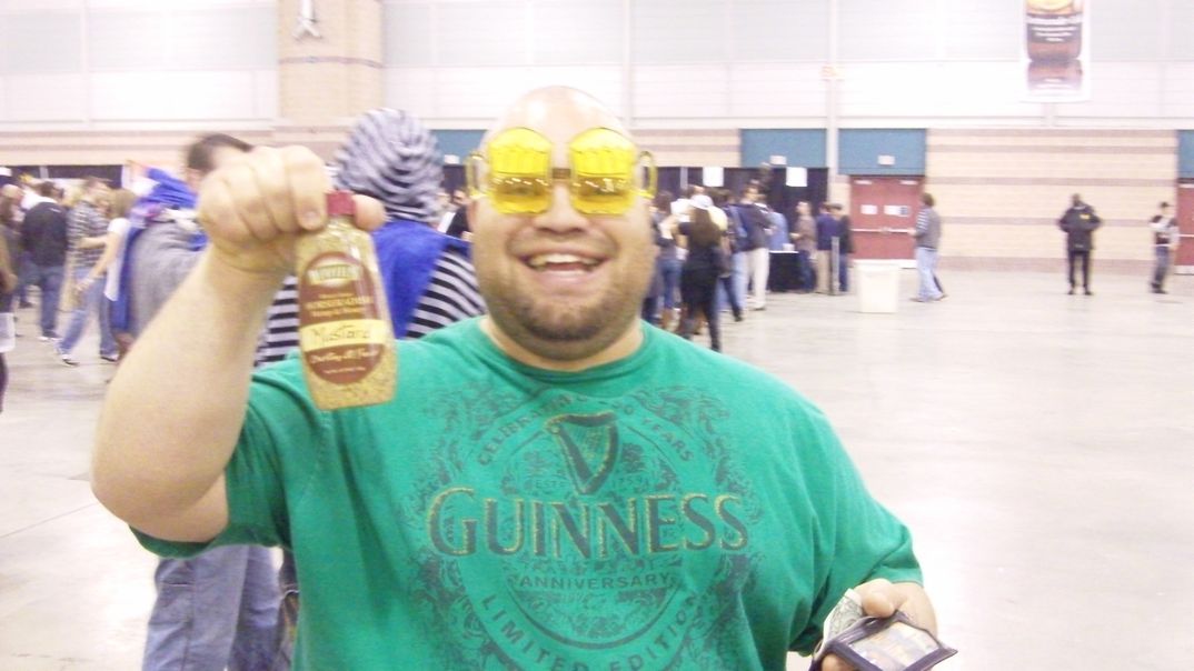 Smiling fan in green short sleeve shirt holding Hempzels(tm) Mustard bottle in convention center with crowds.