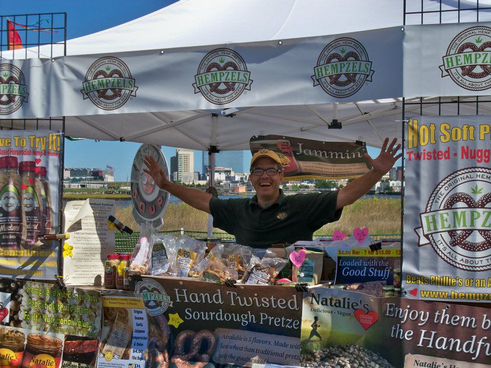 Owner Shawn standing in his booth arms spread wide, big smile wearing his Hempzel(tm) baseball capt with signs and products surrounding him.