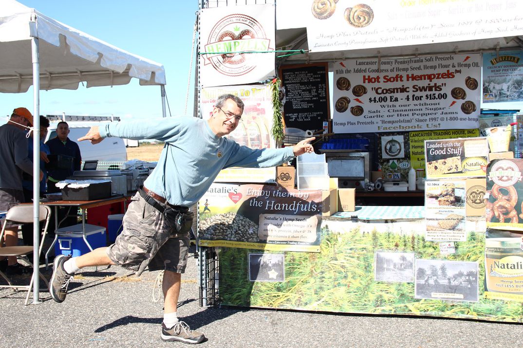 Owner Shawn House posing with left hand pointing & right foot up with booth behind him showing hemp fields, pretzel signs.