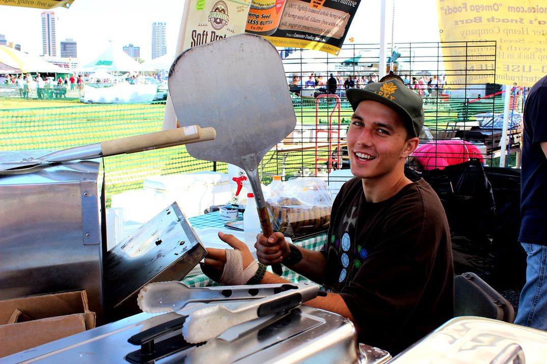 Smiling Philip Holds up a Pretzel Piel in one hand and pretzel oven door in the other tongs in the forefront.