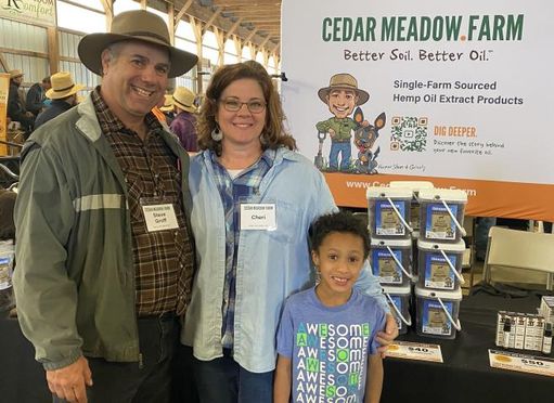 Steve Groff, wife Cheri and grandson stand side by side with amish in background and cedar meadow farms sign in back.